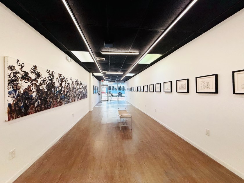 Interior view of an art gallery featuring a long hallway with an abstract painting on one wall and framed artworks on the opposite wall. A single chair is placed in the center, with large windows allowing natural light to enter.